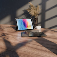 A tablet in a dark gray folio case with an attached keyboard and trackpad sits on a wooden desk, styled with a vase of dried grass and a white cup under warm window shadows.