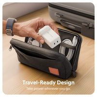 A dark gray zippered travel organizer pouch on a wooden surface, shown open with a hand placing a white travel adapter inside. White cables are stored in the compartments, and a gray suitcase is visible in the background.