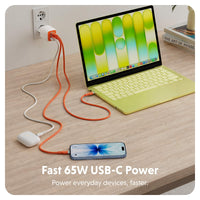 A 65W USB-C power adapter shown in a home office setting, plugging into a wall outlet and connected to both a laptop and a smartphone. The adapter itself is white with an orange cable running from it.